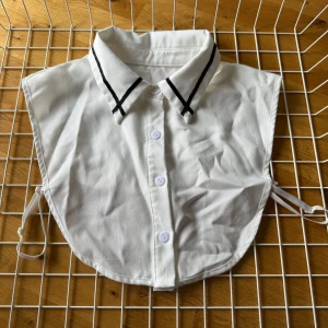 Vit löskrage med svarta detaljer - Säljer en vit löskrage med svarta detaljer. Kragen har knappar framtill och justerbara band på sidorna för att hålla den på plats. Perfekt för att lägga till en stilren touch till vilken outfit som helst. Kragen är i bomull och har en klassisk design med svarta linjer på kragen.