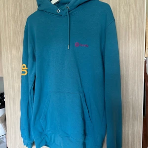 Blå hoodie från Spotify - Säljer en snygg blå hoodie från Spotify. Den har en justerbar huva med dragsko och en stor ficka på magen. På bröstet finns en liten Spotify-logga i rosa och på ena ärmen står det 'SOUNDS UP' i gult. Perfekt för höst och vinter, och den är i nyskick!