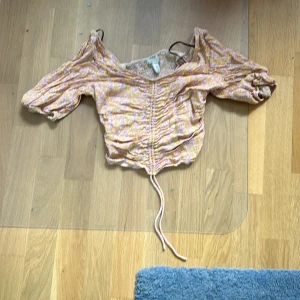 Blommig offshoulder topp - Säljer en söt blommig offshoulder topp i ljusrosa och beige nyanser. Toppen har korta ärmar och en knytning framtill som ger en fin rynkad effekt. Perfekt för sommardagar eller en festlig kväll. Toppen är i bra skick och redo för en ny ägare!