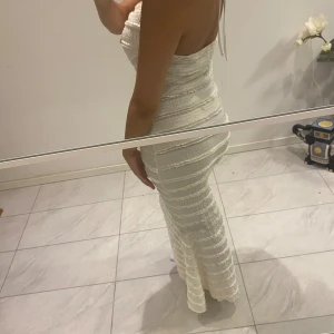 Vit långklänning med ringdetalj - En elegant vit långklänning med en unik ringdetalj framtill. Klänningen har en åtsittande passform och är ärmlös med smala axelband. Materialet har en texturerad yta som ger en intressant visuell effekt.