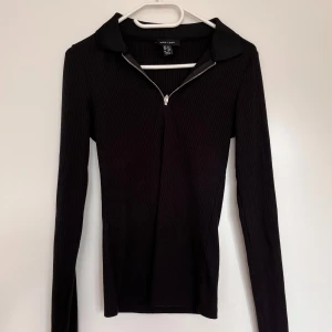 Svart ribbad tröja från New Look - Säljer en snygg svart ribbad tröja från New Look. Den har en dragkedja framtill och en klassisk krage. Tröjan är långärmad och gjord av polyester, viskos och elastan, vilket gör den både bekväm och stretchig. Perfekt för höst och vinter! 🖤