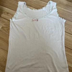 Vit ärmlös topp med spetsdetaljer och rosett - Säljer en söt ljusrosa ärmlös topp med spetsdetaljer runt halsen och ärmhålen. Toppen har en liten rosa rosett framtill som en fin detalj. Perfekt för sommaren eller som ett lager under en kofta. Toppen är i bra skick och väldigt bekväm att ha på sig.