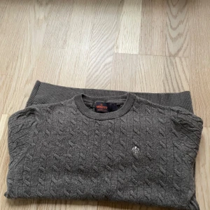 Grå stickad tröja från Morris - Säljer en snygg och bekväm grå stickad tröja från Morris. Tröjan är i storlek L och är tillverkad av 100% ren ull, vilket gör den både varm och mjuk. Den har ett klassiskt flätmönster och en liten broderad logga på bröstet. Perfekt för höst- och vinterdagar!