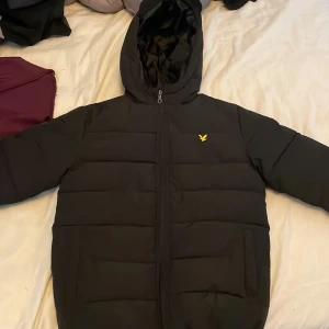 Svart pufferjacka från Lyle & Scott Junior - En svart pufferjacka från Lyle & Scott Junior med huva och dragkedja. Jackan har ett litet gult logomärke på bröstet och är quiltad för extra värme.