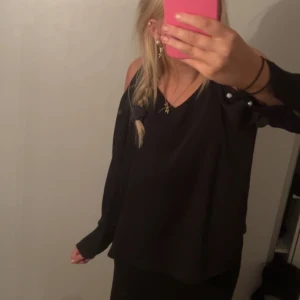 Svart off-shoulder topp - Säljer en snygg svart off-shoulder topp i storlek S. Toppen har en lös passform och är perfekt för festliga tillfällen. Den är ärmlös med en stilren design och hål på axeln som en fin detalj. Passar perfekt till både jeans och kjol!