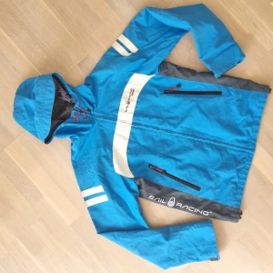 Sail Raicing Windstopper jacka - Windstopperjacka med huva från Sail Raicing, blå med grå, vita och svarta detaljer. Begagnat skick, nötningar finns och gummidetaljerna som täcker kardborren vid ärmsluten har gått av (se bild 2.) 100 % polyester