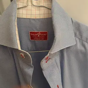 Ljusblå skjorta från Tailor Store Ärmar med dubbel manschett  Små tecken på slitage men fortfarande i gott skick. Skjortan tvättad och pressad på kemtvätt inför försäljningen  Storlek 39 (39cm bred och 75cm lång) 100% bomull