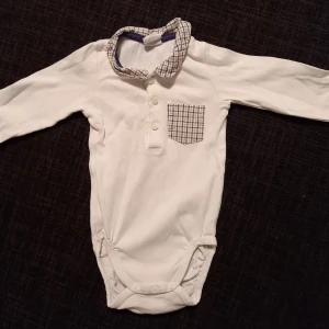 Body med krage strl. 68 - Vit body med rutig ficka, krage och armlappar i storlek 68 från Hm. I fint begagnat skick, inga hål eller fläckar.  Tvättad i parfymfritt tvättmedel och utan sköljmedel.