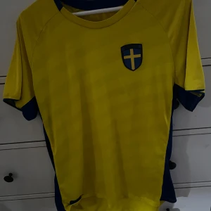 Gul fotbollströja med blå detaljer - En gul fotbollströja med blå detaljer och ett emblem med ett gult kors på en blå sköld på bröstet. Tröjan har korta ärmar och en rund halsringning.