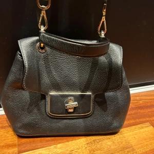 Michael Kors Väska - En axelbandsväska som har varit en favorit i garderoben, därav ser den använd ut varav de slitningsskador som visas på bild.  Mått:  Bredd: 21-29 cm Höjd: 19 cm Djup: 9cm längst ner, 4 cm högst upp (som kan knäppas upp och bli 9cm)