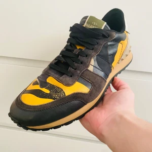 Valentino sneakers med kamouflagemönster - Ett par Valentino sneakers med ett unikt kamouflagemönster i svart, gult och beige. Skorna har svarta snören och en beige sula. De är i välanvänt skick.