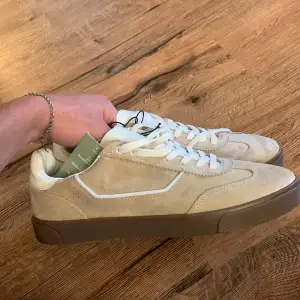 Ett par helt nya beige sneakers från H&M i mocka med vita snören och en brun sula. Skorna har en klassisk design med en bekväm passform. Säljer då de inte passade