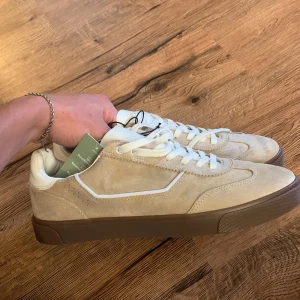 Beige sneakers från H&M - Ett par helt nya beige sneakers från H&M i mocka med vita snören och en brun sula. Skorna har en klassisk design med en bekväm passform. Säljer då de inte passade