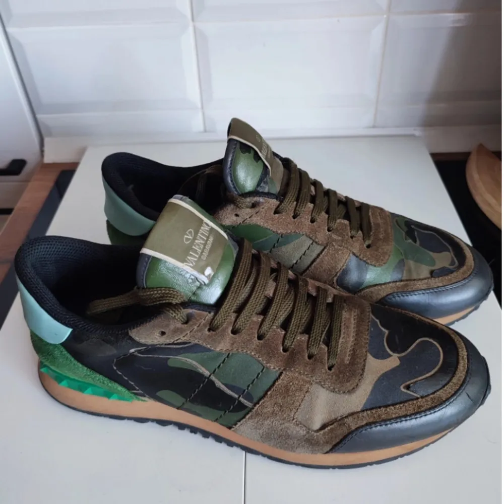 Ett par Valentino Rockrunner sneakers i camouflagefärger med gröna, bruna och svarta detaljer. Skorna har en kombination av mocka och skinnmaterial samt en gummisula. De har snörning framtill och en unik design med nitar på hälen.. Skor.