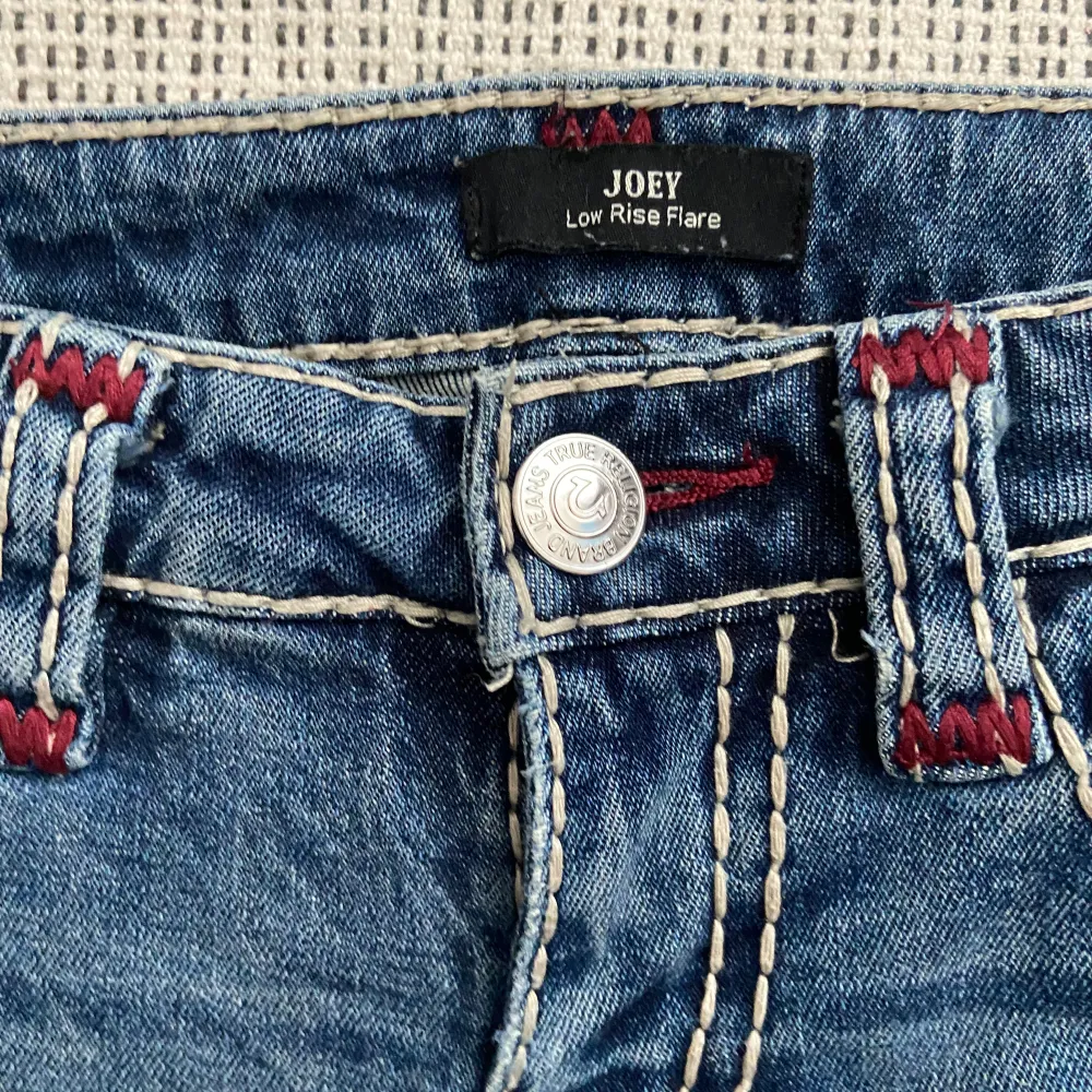 Blå jeansbyxor från True Religion med låg midja och utsvängda ben. Byxorna har dekorativa sömmar i rött och blått samt fickor med knappar på baksidan. De är något slitna vid bensluten.. Jeans & Byxor.