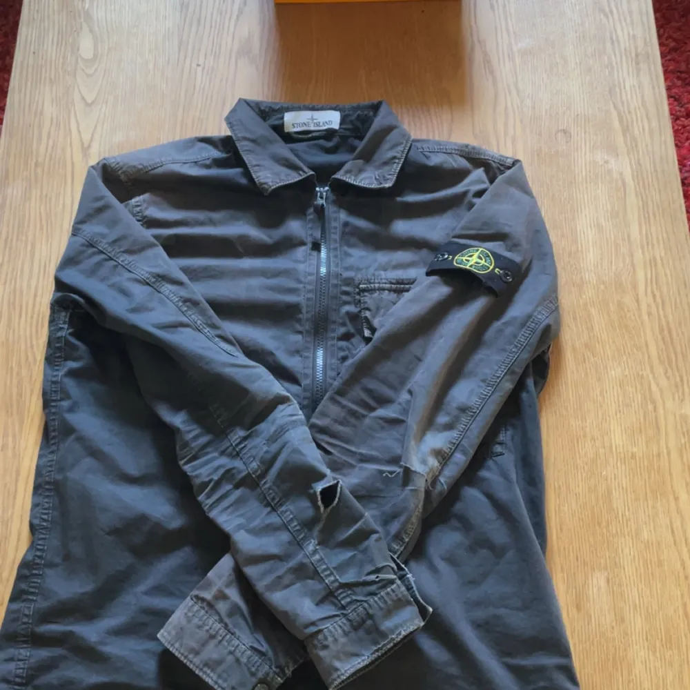 En grå overshirt från Stone Island med dragkedja framtill och en bröstficka med dragkedja. Jackan har en krage och en logotyp på vänster ärm. Den är i välanvänt skick med synliga slitningar och hål på ärmarna.. Jackor.