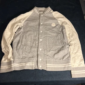 Gråvit moncler tröja  - En hög kvalite gråvit collegetröja av moncler gjord i bomull med vita metallknappar. Perfekt till en frisk vårdag, höstdag eller sommarkväll om man vill se lite extra snygg ut!