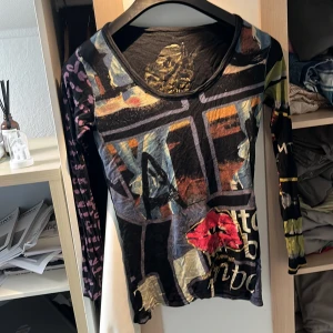 Flerfärgad långärmad tröja med tryck - En unik och färgglad långärmad tröja med olika tryck och mönster. Tröjan har en rund halsringning och är tillverkad i ett tunt material. Den har en blandning av färger som svart, röd, gul, blå och grön med olika grafiska element och text.