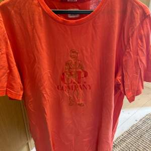 mycket bra skick, knappt använd. En orange t-shirt från C.P. Company med ett tryck av en figur och texten 'C.P. Company' på framsidan. T-shirten har korta ärmar och en rund halsringning. 