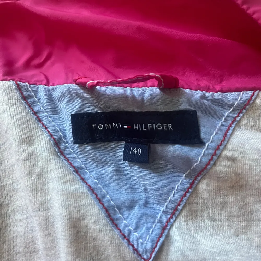 En rosa jacka från Tommy Hilfiger med dragkedja framtill. Jackan har långa ärmar och är i ett lätt material, vilket gör den perfekt för vår- och höstsäsonger.. Takit.