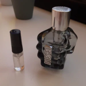 Diesel Only The Brave parfym sample - En parfymflaska från Diesel formad som en knuten näve med en silverfärgad kork. Flaskan är genomskinlig med en mörk toning och har Diesel-logotypen på framsidan.