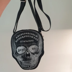 Banned ouija skull väska - Cool axelväska från banned köpt för flera år sen men använd max 5 gånger, ett stort fack med 2 små fack inuti. Inga slitningar på fake lädret.