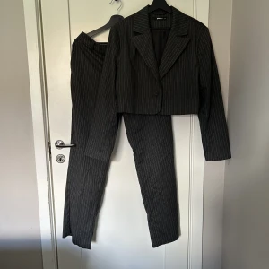 Kostymset  - Det med kostymbyxor och Croppad kavaj från Ginatricot. Superfint och i nyskick. Grårandigt mönster och mjukt material. Överdelen är storlek 40 och underdelen 42. SE MIN PROFIL för referensmått. 