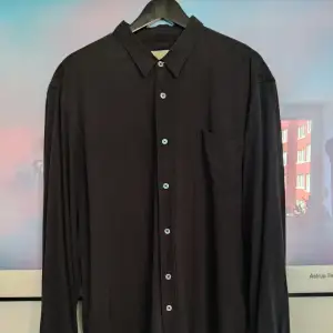 Our Legacys klassiska skjorta i svart. Avslappnad passform. 100% kinesiskt råsilke. Knappar i pärlemor. Använd flera gånger, men utan skador eller slitningar.  https://www.ourlegacy.com/classic-shirt-black-silk-core