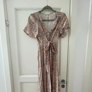 Sommarklänning  - Lång blommig sommarklänning med knyte och knappar i midjan. Aldrig använd, helt nytt skick. Fin och färgglad maxi klänning. Perfekt under vår och sommar. 100%viskos köpt för ca500kr