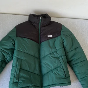 North Face jacka - En varm och bekväm pufferjacka från The North Face i grönt och svart. Jackan har en dragkedja framtill och är quiltad för extra värmeisolering. Perfekt för kalla vinterdagar. Använt i en vinter
