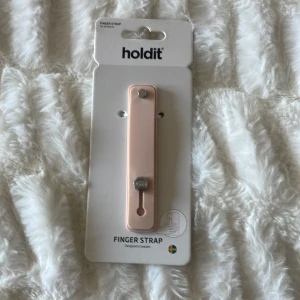 Finger Strap från Holdit - En fingerrem från Holdit i en ljusrosa färg. Remmen är designad för att fästa på baksidan av en mobiltelefon för att ge ett bättre grepp. passar till alla mobiler 