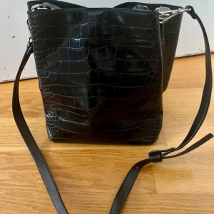 Svart väska - Svart väska i skinnimitation. Långt avtagbart band samt ett kortare handtag. Tre fack, ett med dragkedja. Stängning med knapp. Nyskick utan prislapp. 