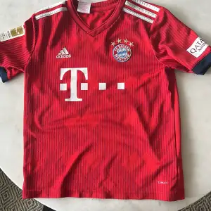 En röd Bayern München hemmatröja från säsongen 2017/1018 . Skicket är okej, på framsidan är det fler små knottar av tyg som uppstått efter man tvättat tröjan. Men man märker knappt av dessa. Storleken är barn M. 