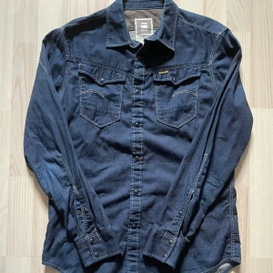 Mörkblå G-stars skjorta - 149kr - Mörk blå G-star raw skjorta i storlek large. 100% bomull. Ljus söm, svart & gul ”G-STAR” broderad tag på vänstra bröstfickan. För mått/fler bilder skriv privat.
