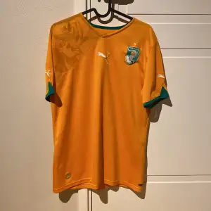 En Elfenbenskusten fotbollströja från säsongen 2010/2011 säljes. Tröjan har en attraktiv design, är i okej skick och passar för fans av landslaget eller fotbollssamlare. En unik tröja med fin historia.