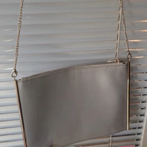 Grå axelväska med kedja - En elegant grå axelväska med guldfärgade detaljer och en kedja som axelrem. Väskan har en enkel och stilren design med en dragkedja på insidan för extra förvaring.från Dorothy Perkins i mycket bra skick. Behöver rengöring i fickan på väskan på grund av smink, se bilder. Kan användas utan eller med förlängningsbälte.