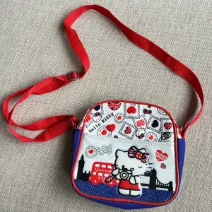 Söt liten väska med motiv av Hello Kitty Globetrotter collection -UK  Färger röd, vit och blå. Justerbar axelrem. Storlek ca 17x16 cm. Ett fack som stängs med dragkedja. 
