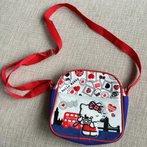 Väska Hello Kitty - Söt liten väska med motiv av Hello Kitty Globetrotter collection -UK  Färger röd, vit och blå. Justerbar axelrem. Storlek ca 17x16 cm. Ett fack som stängs med dragkedja. 