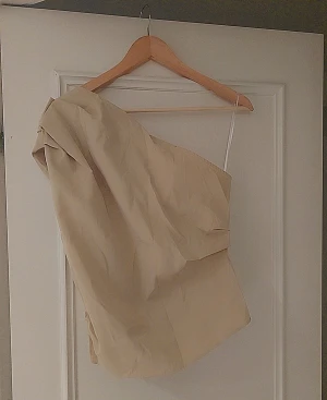 Beige oneshoulder topp från H&M - Säljer en beige oneshoulder topp från H&M  storlek M. Toppen har en cool asymmetrisk design med en axelbandslös sida och ett smalt axelband på andra sidan. Materialet är lätt och känns som bomull, perfekt för att sticka ut med en unik siluett.