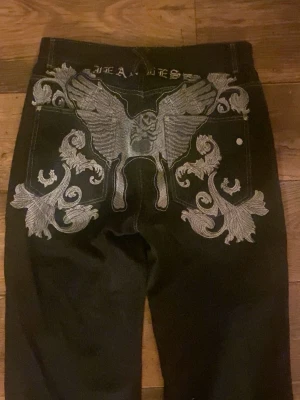 Svarta jeans med silverbroderi - Unika svarta jeans med stora silverbroderade detaljer och vingar på bakfickorna. Sitter skit snyggt baggy på mig som är 163cm. coola broderier även framtill. Lite mer tunt material, sitter också bra i midjan på mig som brukar ha s. Slitna längst ner