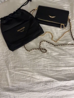 Svart axelväska från Zadig & Voltaire - Snygg svart axelväska i skinn från Zadig & Voltaire med guldfärgad kedjerem och detaljer. Väskan har dragkedja både fram och bak samt ikonisk vinglogga i guld på framsidan. Perfekt storlek för det viktigaste och ger en cool vibe till din outfit.