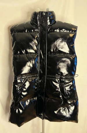 Svart dunväst från H&M - Svart dunväst från H&M i storlek S med glansig finish och hög krage. Västen har dragkedja och tryckknappar framtill, två fickor och snörning i midjan för att justera passformen. Perfekt för att ge din outfit en cool edge.
