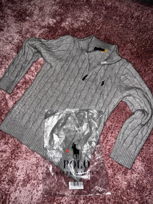 Grå stickad half zip från Polo Ralph Lauren - Säljer en grå stickad tröja från Polo Ralph Lauren med half zip och kabelstickat mönster. Tröjan har långärmade ribbade muddar och klassisk krage. Perfekt för dig som gillar stilren och tidlös design med snyggt broderad logga på bröstet.