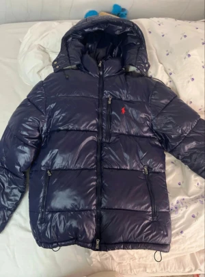 Marinblå glänsig pufferjacka från Polo Ralph Lauren - Säljer en marinblå pufferjacka från Polo Ralph Lauren med blank finish och röd broderad logga på bröstet. Jackan har huva, dragkedja framtill och elastiska muddar. Perfekt för kalla dagar och riktigt snygg streetstil. Pris går att diskutera! 💯🫡