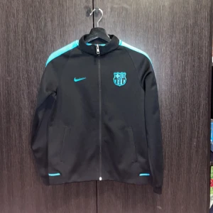 Svart Nike FC Barcelona  - Svarttröja  från Nike med FC Barcelona-logga och turkosa detaljer på axlar, ärmar och bröst. Jackan har hel dragkedja framtill, ståkrage och två sidofickor. Perfekt för dig som älskar fotboll och vill ha en sportig look.
