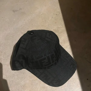 Svart keps med logomönster - Snygg svart keps med diskret FF-logomönster över hela kepsen. Justerbar rem baktill i svart läder med Fendi-tryck. Kepsen är tillverkad i ett tygmaterial och har klassisk böjd skärm. Perfekt för dig som gillar exklusiva accessoarer.