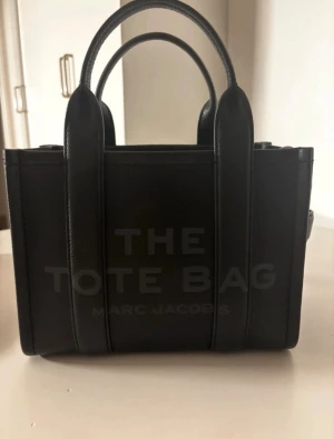 Svart The Tote Bag från Marc Jacobs - Säljer min Marc Jacobs Tote Bag i svart äkta läder då jag tyvärr inte helt är nöjd med storleken. Väskan är i princip helt ny, använd endast 2 gånger. Inga defekter, repor eller märken, den ser ut som ny.  100% äkta autenticitetspapper och kort medföljer  Material: 100% ko-läder Färg: Svart  Modell: The Tote Bag