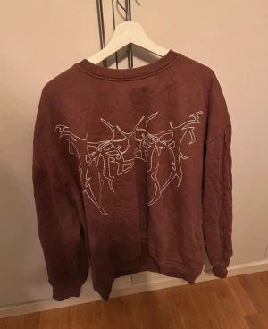 Brun sweatshirt med tribaltryck - Säljer en brun oversized sweatshirt med ett fett vitt tribaltryck på framsidan. Tröjan har rund hals och långa ärmar, perfekt för dig som gillar streetstyle och vill sticka ut lite extra. Materialet känns mjukt och skönt mot huden.