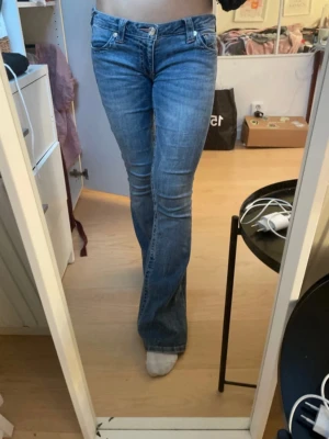 Blå bootcut jeans från True Religion - Säljer ett par blå jeans från True Religion med klassisk bootcut och låg midja. Jeansen har slitna detaljer vid bensluten, fem fickor och synliga sömmar. Materialet är jeans med en mjuk känsla och passformen är slim upptill med utsvängda ben.