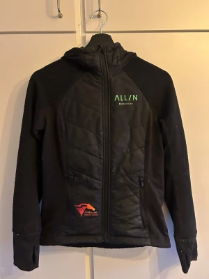 Svart quiltad jacka All In Equestrian - Svart jacka med huva från All In Equestrian, quiltad framsida och rygg, grönt tryck på bröst och rygg. Snygga detaljer med dragkedja och ribbade ärmar. Perfekt för dig som gillar ridning och vill ha en sportig look.